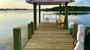 Un muelle de madera que lleva a un pabellón cubierto con una mesa y banca, con vista a agua tranquila y barcos a lo lejos.