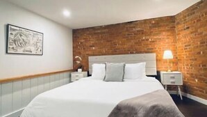 Un dormitorio con una cama grande, ropa de cama blanca, un muro de ladrillo decorativo, una mesita de noche con una lámpara y un cuadro en la pared.