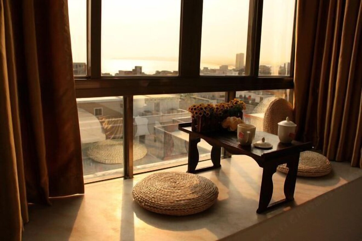 A cozy room with a large window, a small table with a vase of flowers, and two woven poufs.