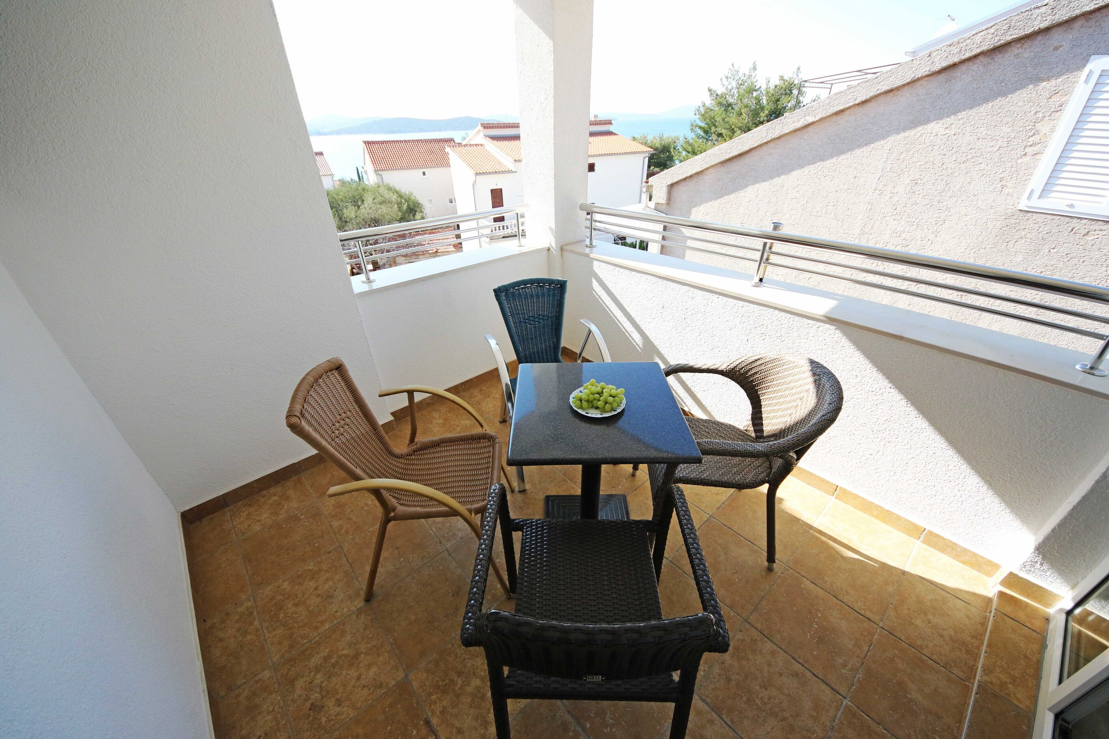 Een balkon met een tafel en stoelen, een schaal fruit en uitzicht op gebouwen en de zee.