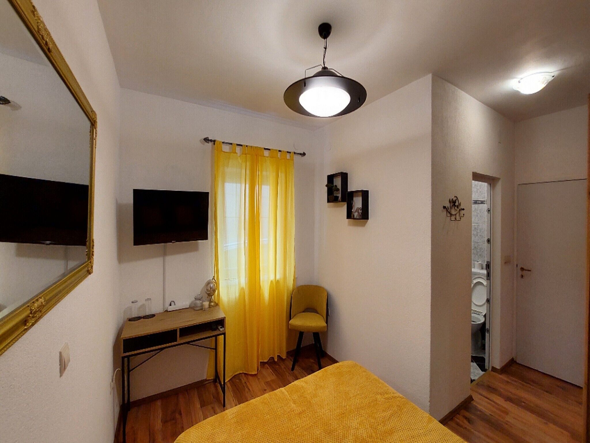 A small, well-lit room with a bed, a desk, a chair, a television, and a door leading to a bathroom.