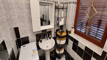 A bathroom with a washing machine, a sink, a mirror, and a window with blinds.