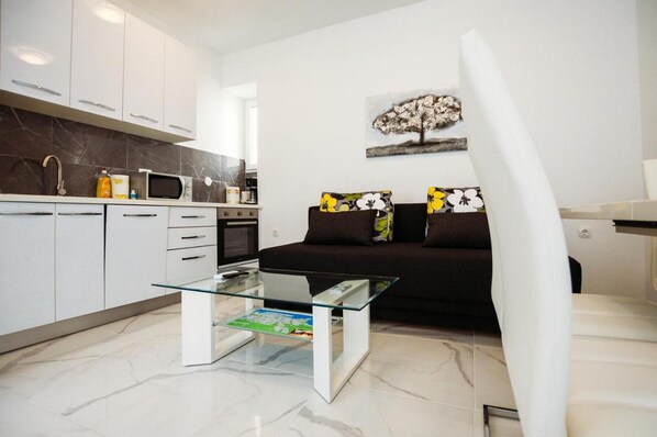 A modern kitchen with white cabinets, a microwave, and a coffee machine. There is a glass coffee table with a map underneath. A dark sofa with floral cushions is against the wall.