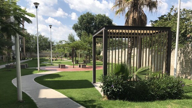 Ein Weg führt zu einem überdachten Sitzbereich mit einer Pergola, umgeben von gepflegtem Rasen und Pflanzen.