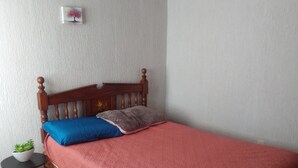 Ein Einzelbett mit Holz-Kopfteil, einer roten Bettdecke und zwei Kissen (ein blaues, ein graues). Neben dem Bett steht ein kleiner runder Tisch mit einem Blumentopf.