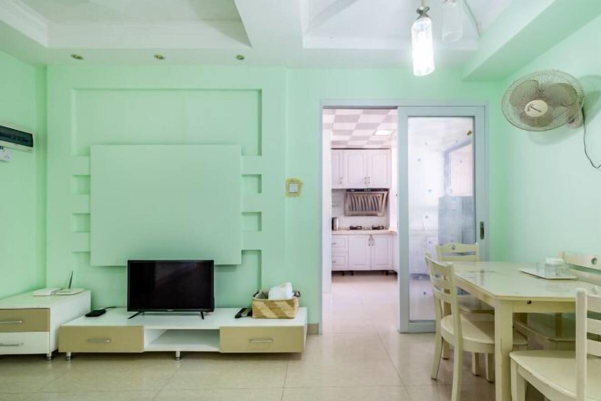 Une pièce avec un téléviseur à écran plat posé sur un meuble blanc, une table à manger avec des chaises, un petit meuble blanc et un ventilateur fixé au mur.