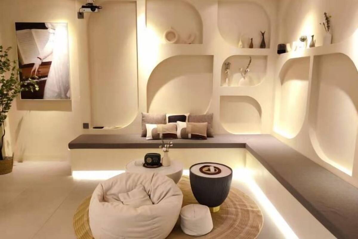 Une pièce moderne et minimaliste, avec un canapé, un fauteuil en forme de poire et une table ronde. On y trouve des étagères ornées d’objets décoratifs et un tableau encadré accroché au mur.