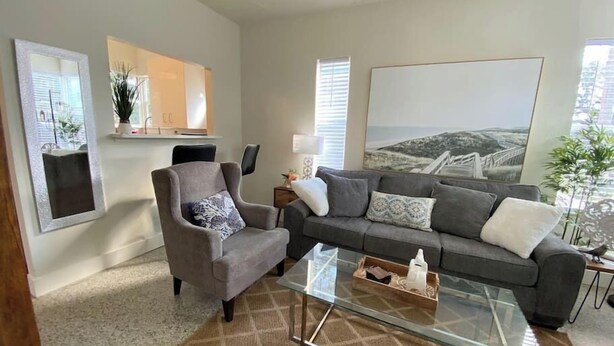 Una sala de estar moderna con un sillón gris, un sofá gris y una mesa de centro de vidrio. Se aprecia un ventanal con persianas, un cuadro en la pared y una planta en el alféizar.