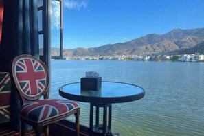 A table and chair by a window with a view of a lake and mountains.