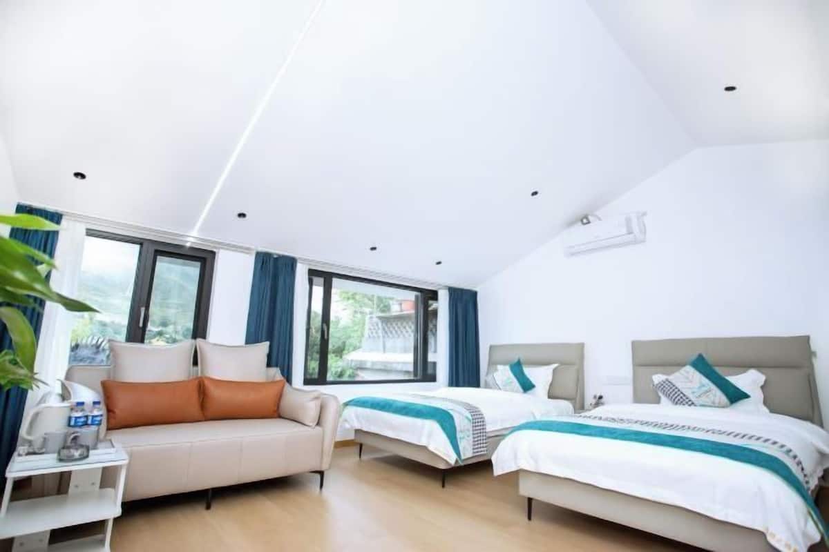 Ett modernt hotellrum med två sängar, en soffa, ett litet bord med en lampa och ett stort fönster med utsikt över grönområden utanför.