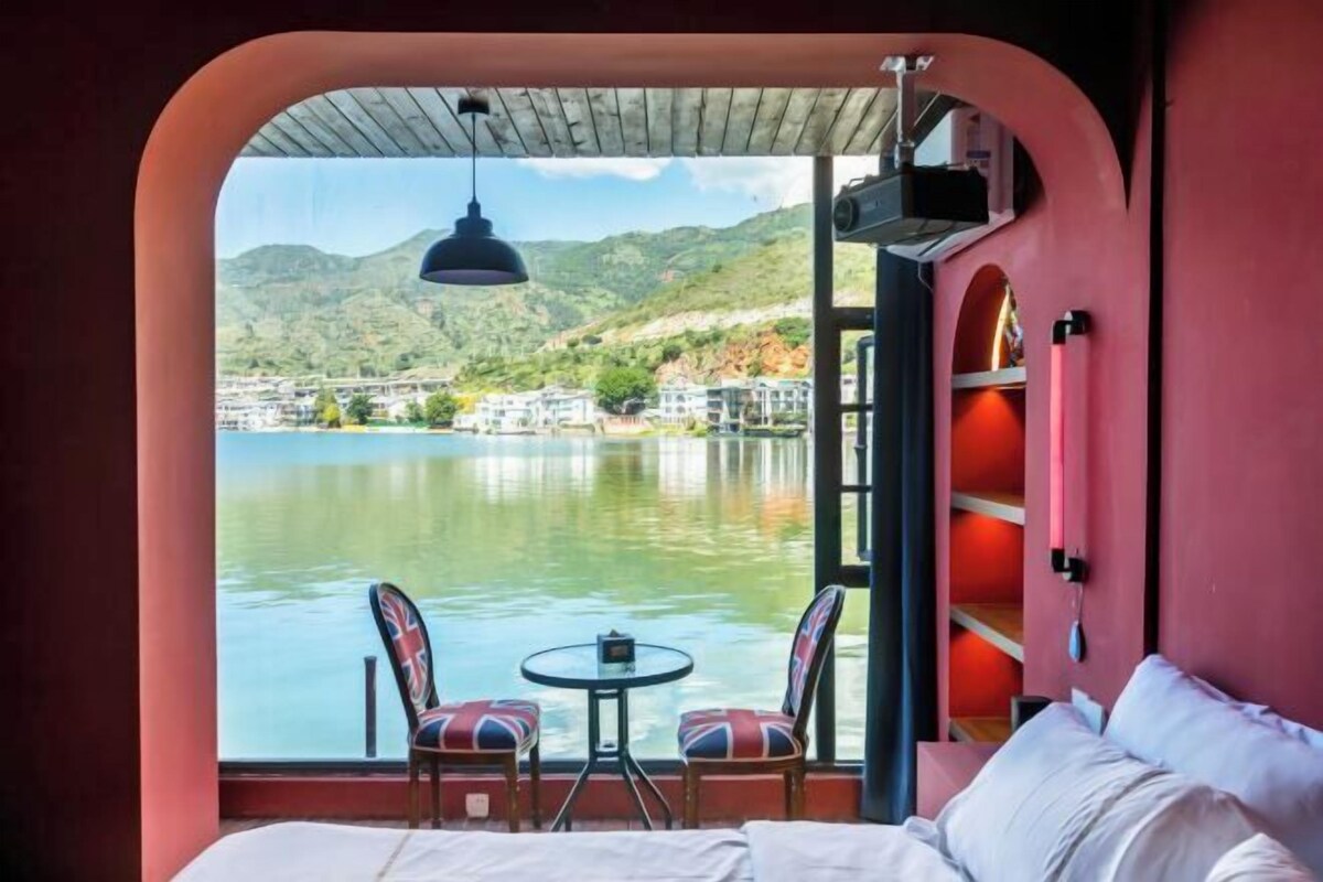A room with a bed, a table with chairs, a view of a lake, and a mountainous landscape.