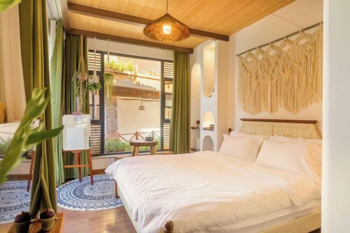 A bedroom with a bed, a hanging light fixture, a window with curtains, and a view of the outside.