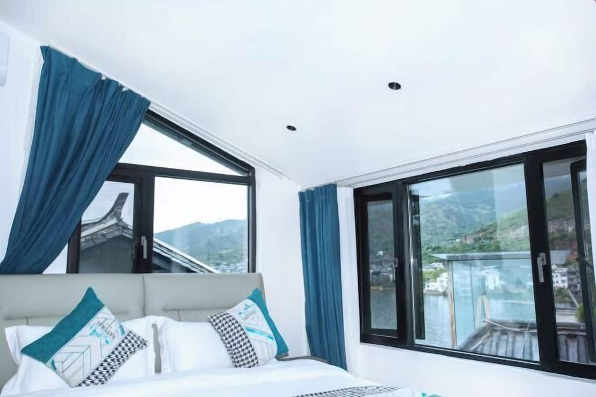 Ett modernt hotellrum med en stor säng, två fönster med gardiner och utsikt över ett bergigt landskap.