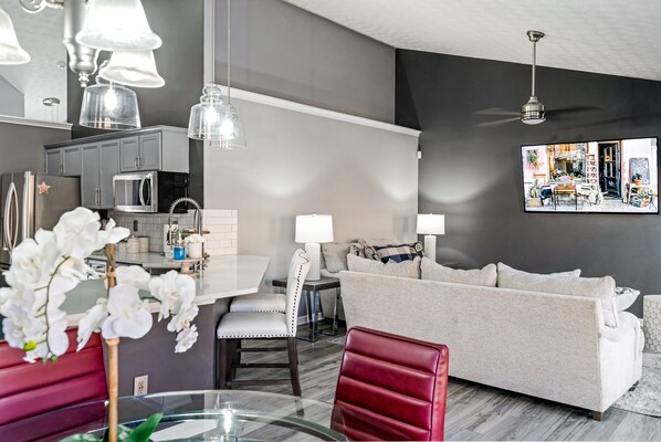 A modern kitchen with grey cabinets, a microwave, and a dining area with a glass table and red chairs. There is a grey sofa and a red chair in the living area. - Enjoyable/Spacious 3bd Farmhouse (Lawrenceville)