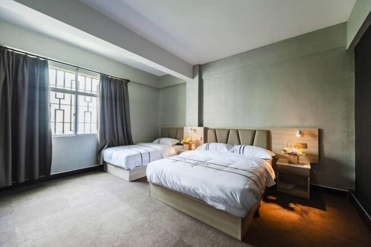 Une chambre d’hôtel moderne avec deux lits, une grande fenêtre avec des rideaux, une table de chevet avec une lampe et un luminaire fixé au mur.