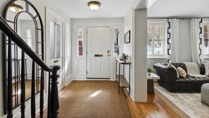 A spacious living room with a large black leather sofa, a wooden floor, and a view of the outside through a window with curtains. - Your Beautiful Savannah House: Picturesque Inside and Out! Walk to Everything (Savannah)