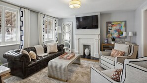 A living room with a leather sofa, a striped ottoman, a white armchair, a fireplace, and a flat-screen TV. - Your Beautiful Savannah House: Picturesque Inside and Out! Walk to Everything (Savannah)