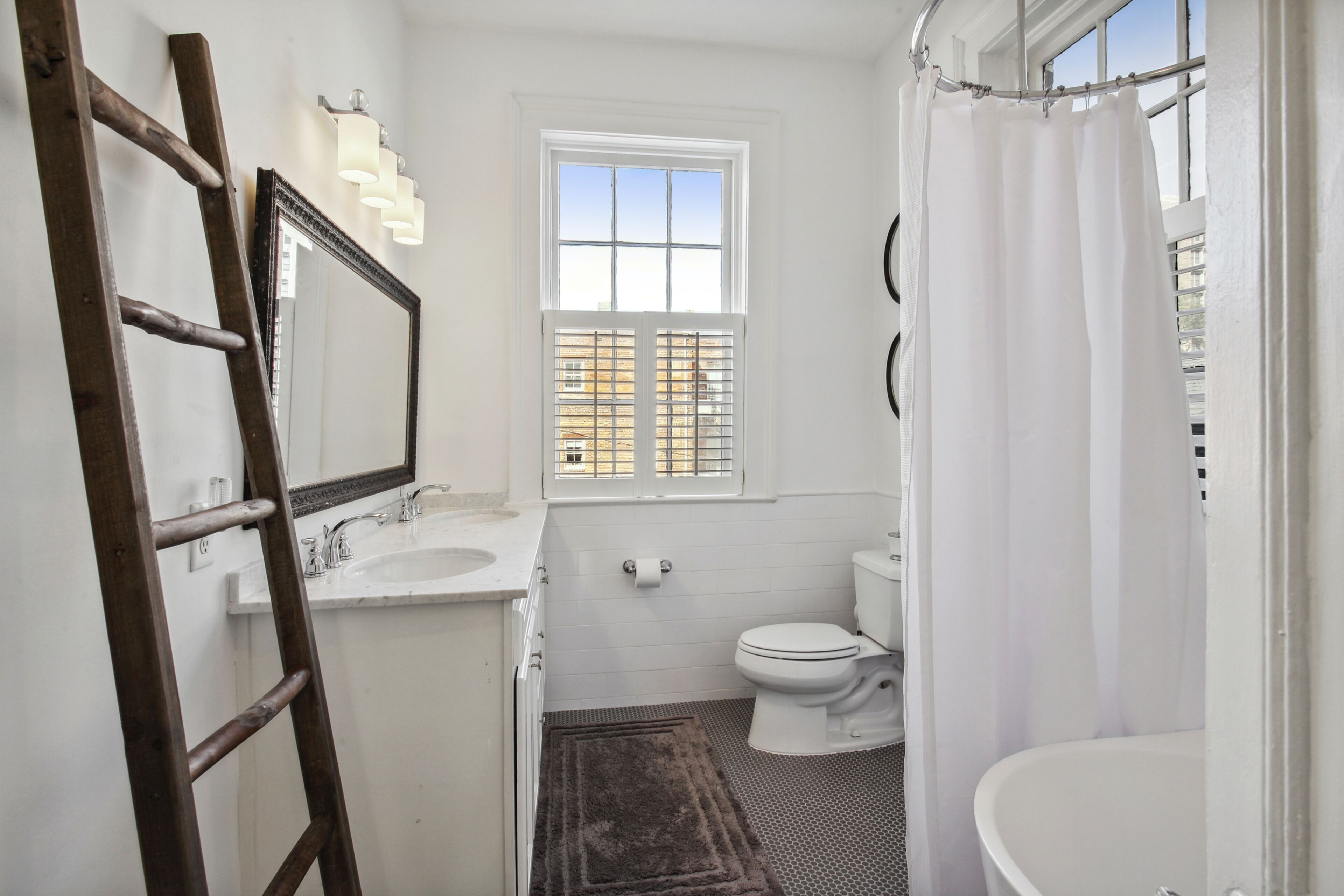 Une salle de bain avec un lavabo, des toilettes et une baignoire, tous de couleur blanche. Il y a une fenêtre au-dessus des toilettes et une échelle appuyée contre le mur.