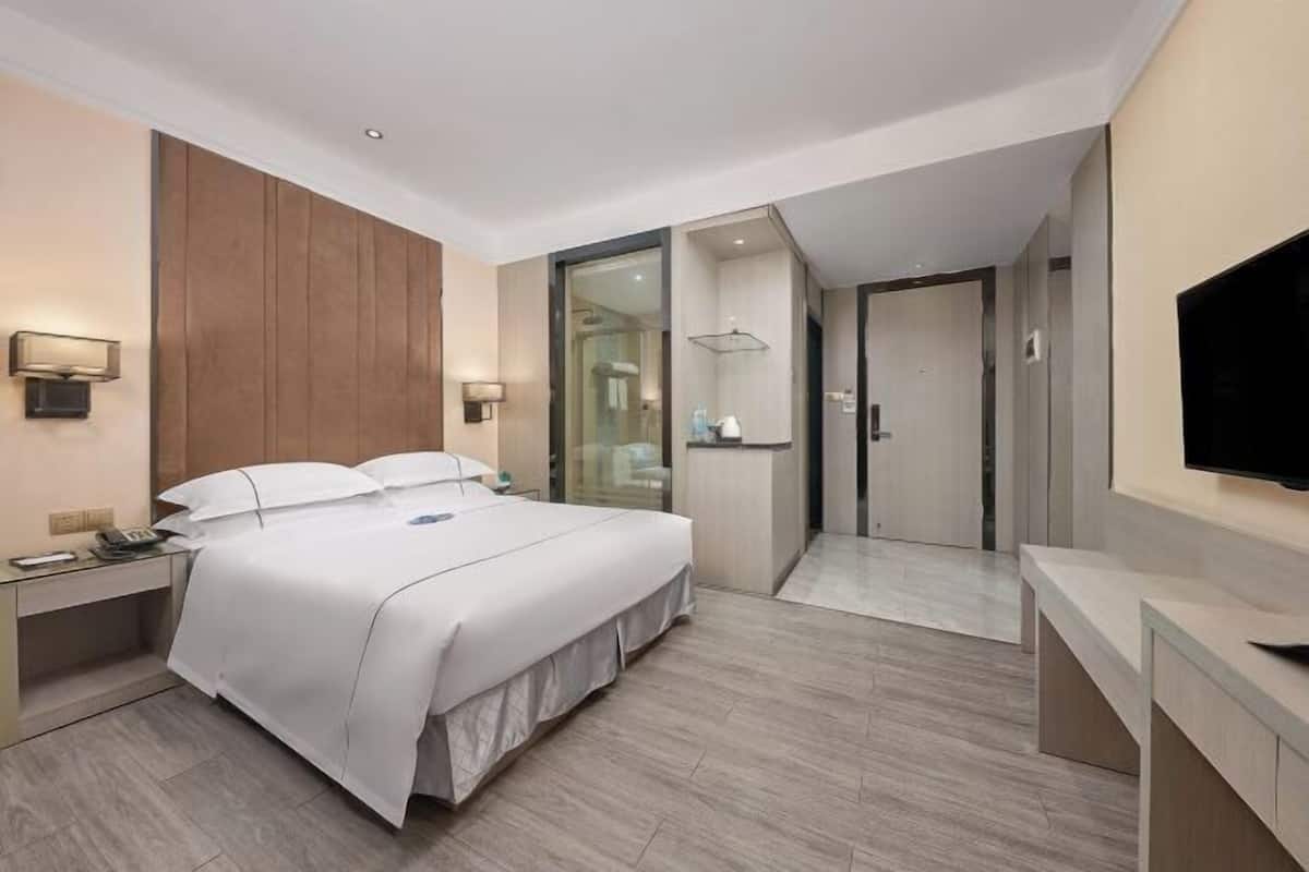 A modern hotel room with a large bed, a desk, a TV, and a bathroom with a glass partition.
