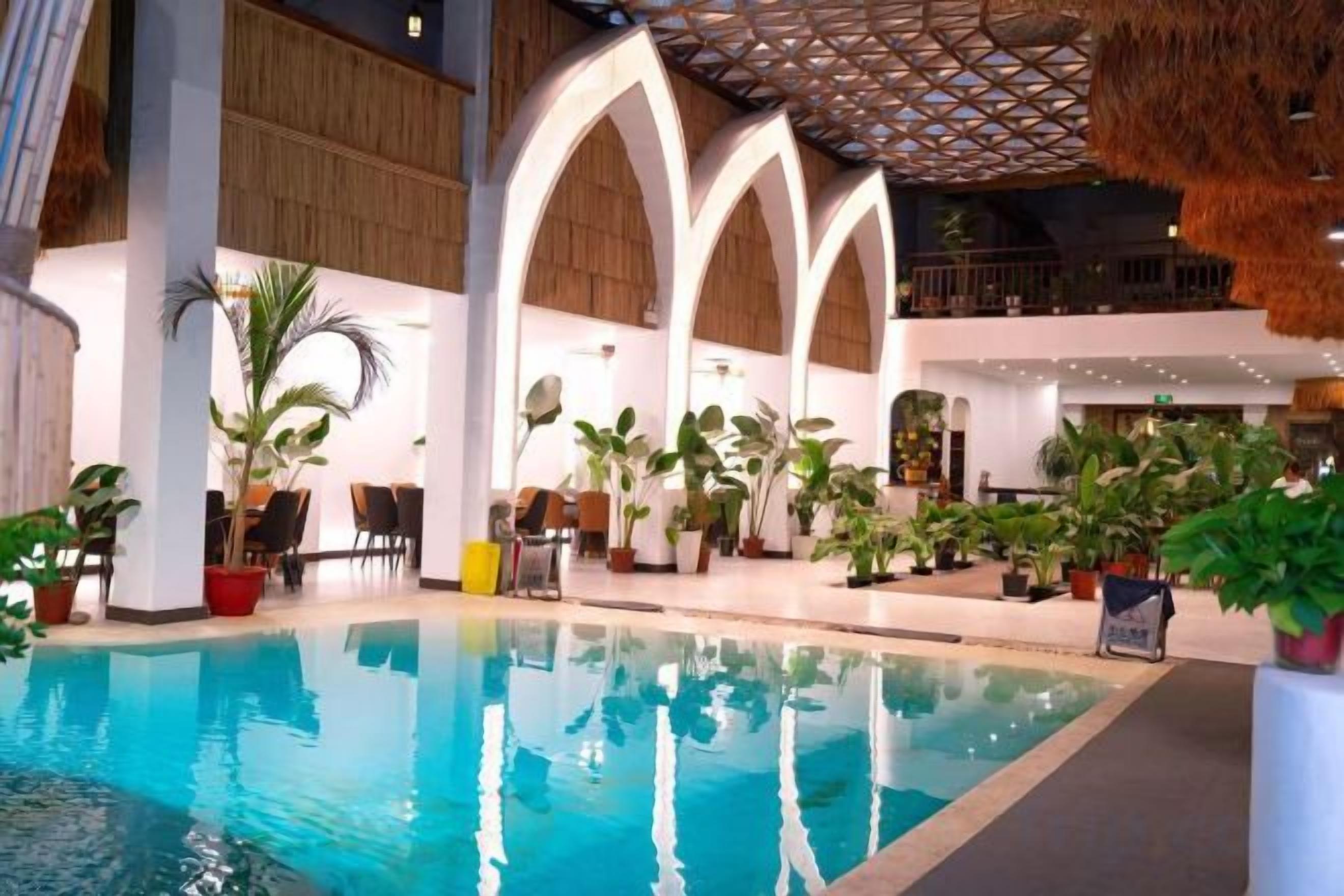 An indoor swimming pool with a thatched roof, surrounded by potted plants and a wooden floor.