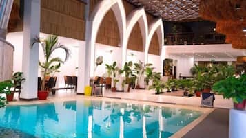 An indoor swimming pool with a thatched roof, surrounded by potted plants and a wooden floor.