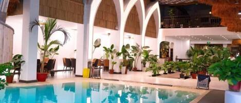 An indoor swimming pool with a thatched roof, surrounded by potted plants and a wooden floor.