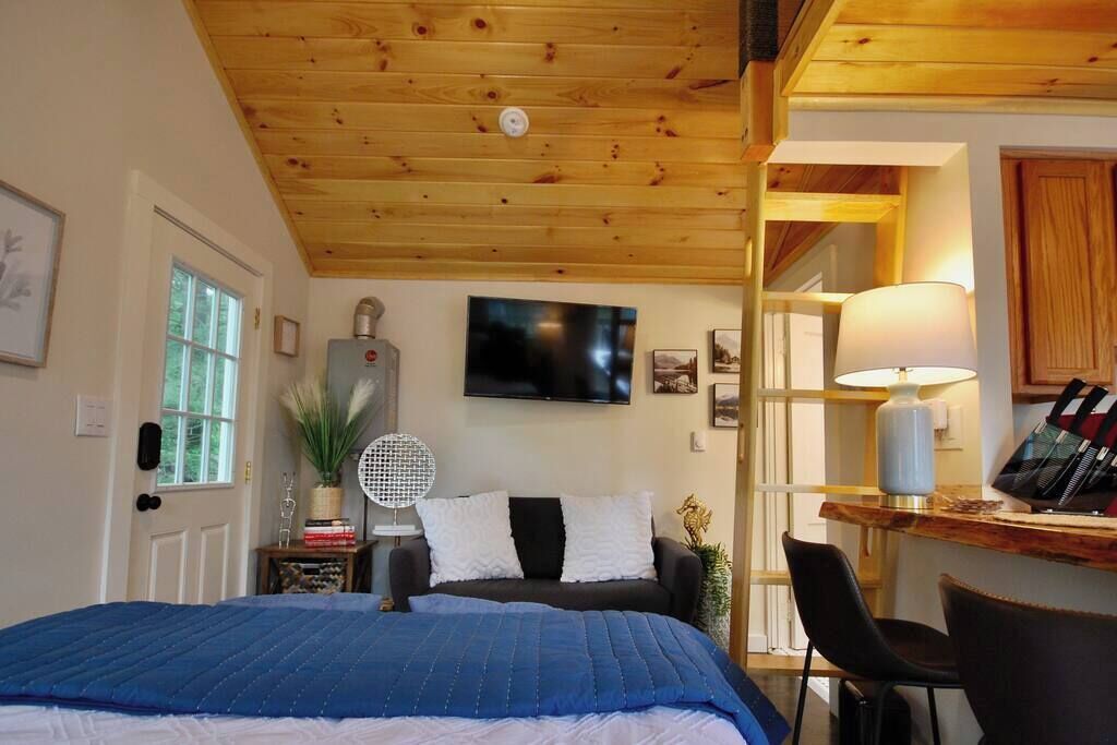 A bedroom with a wooden ceiling, a bed with a blue quilt, a desk with a lamp, and a TV on the wall.
