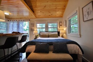 A cozy room with a wooden ceiling, a bed with a blue quilt, two bedside tables with lamps, and a window with a view of trees. - Cheerful Cottage with Loft Close to Everything 6 (Edgecomb)