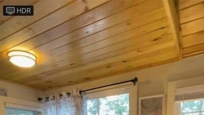 Una habitación acogedora con techo de madera, una pequeña zona de bar con mostrador y taburetes, una ventana con cortinas y una lámpara sobre una mesita.