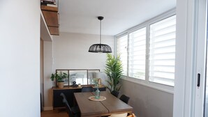 Un comedor con una mesa de madera, sillas, una lámpara colgante, una planta en maceta y una ventana con persianas.