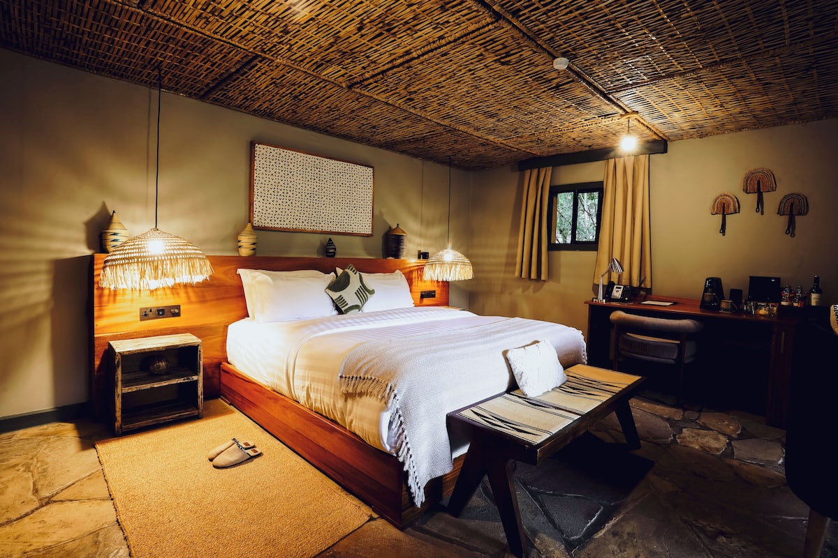 A cozy bedroom with a wooden bed, bedside lamps, a small nightstand, a desk with a chair, and a window with curtains.