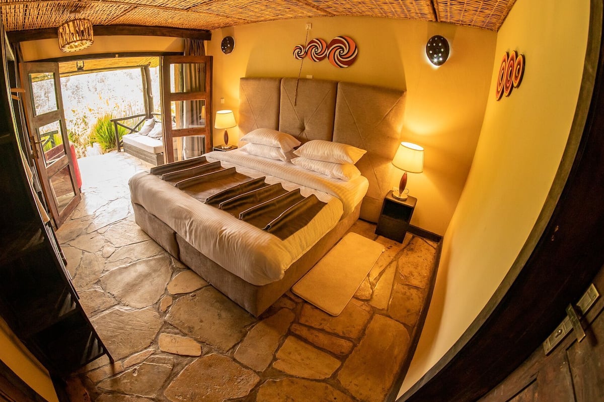 A bedroom with a stone floor, a bed with multiple pillows, a nightstand, and a lamp.
