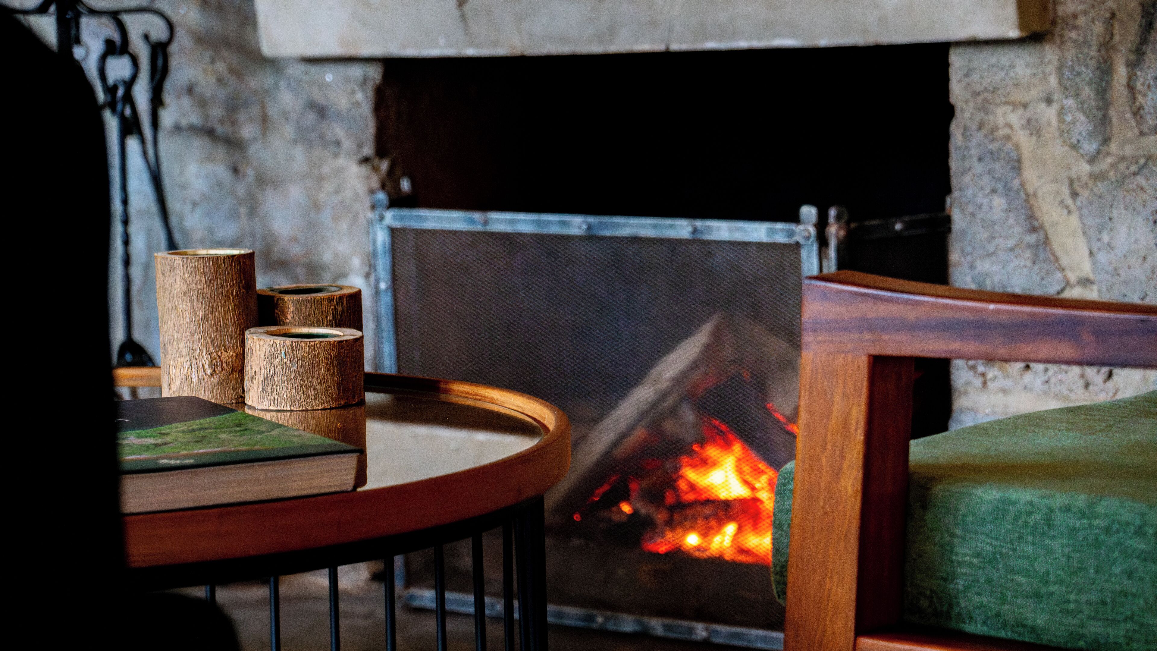 Un rincón acogedor con una mesa redonda de madera, un banco verde acolchado y una chimenea de piedra con fuego visible.