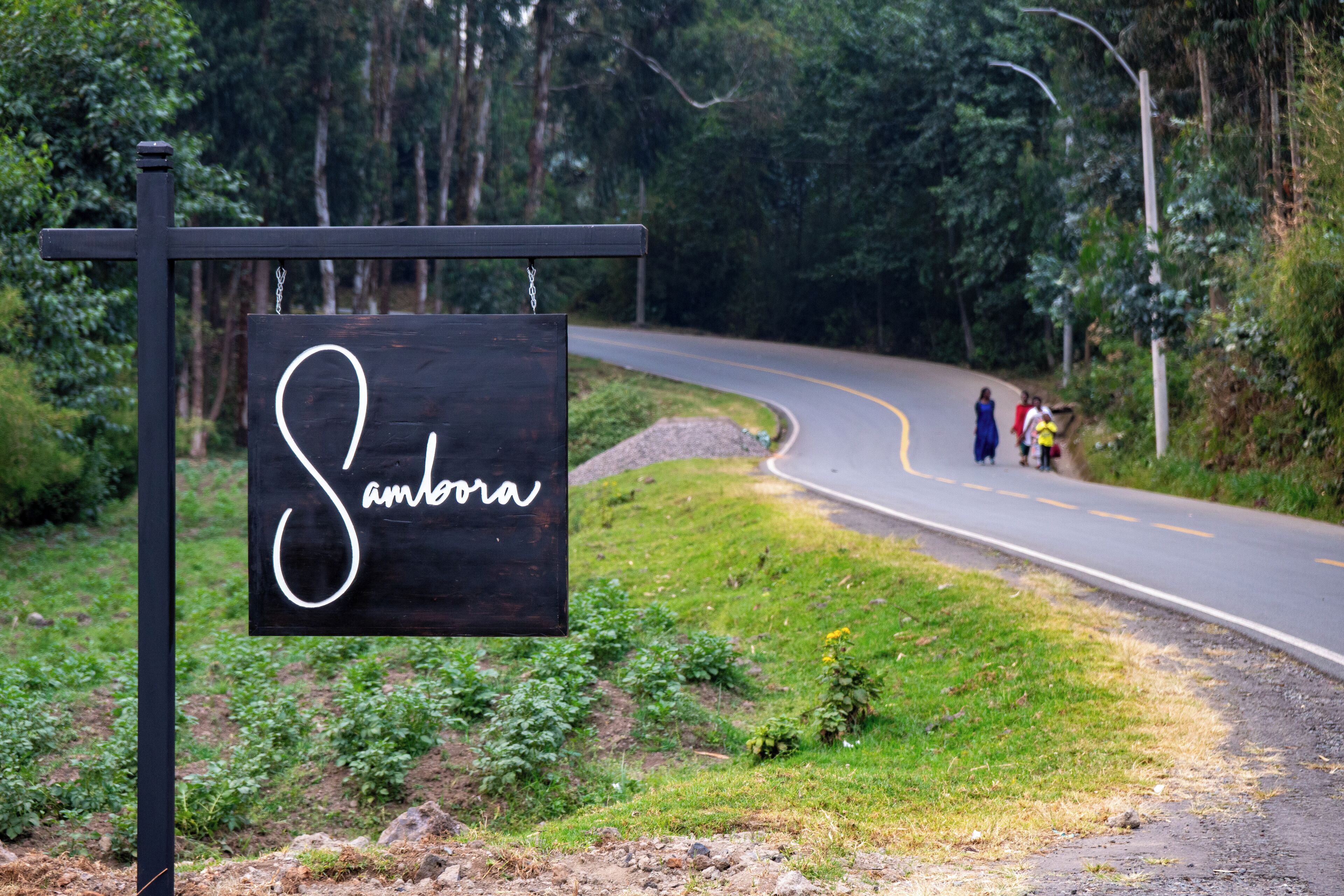 A sign with the name "Sambora" in cursive on a black background, mounted on a metal post beside a curving road with greenery and a few people in the distance.