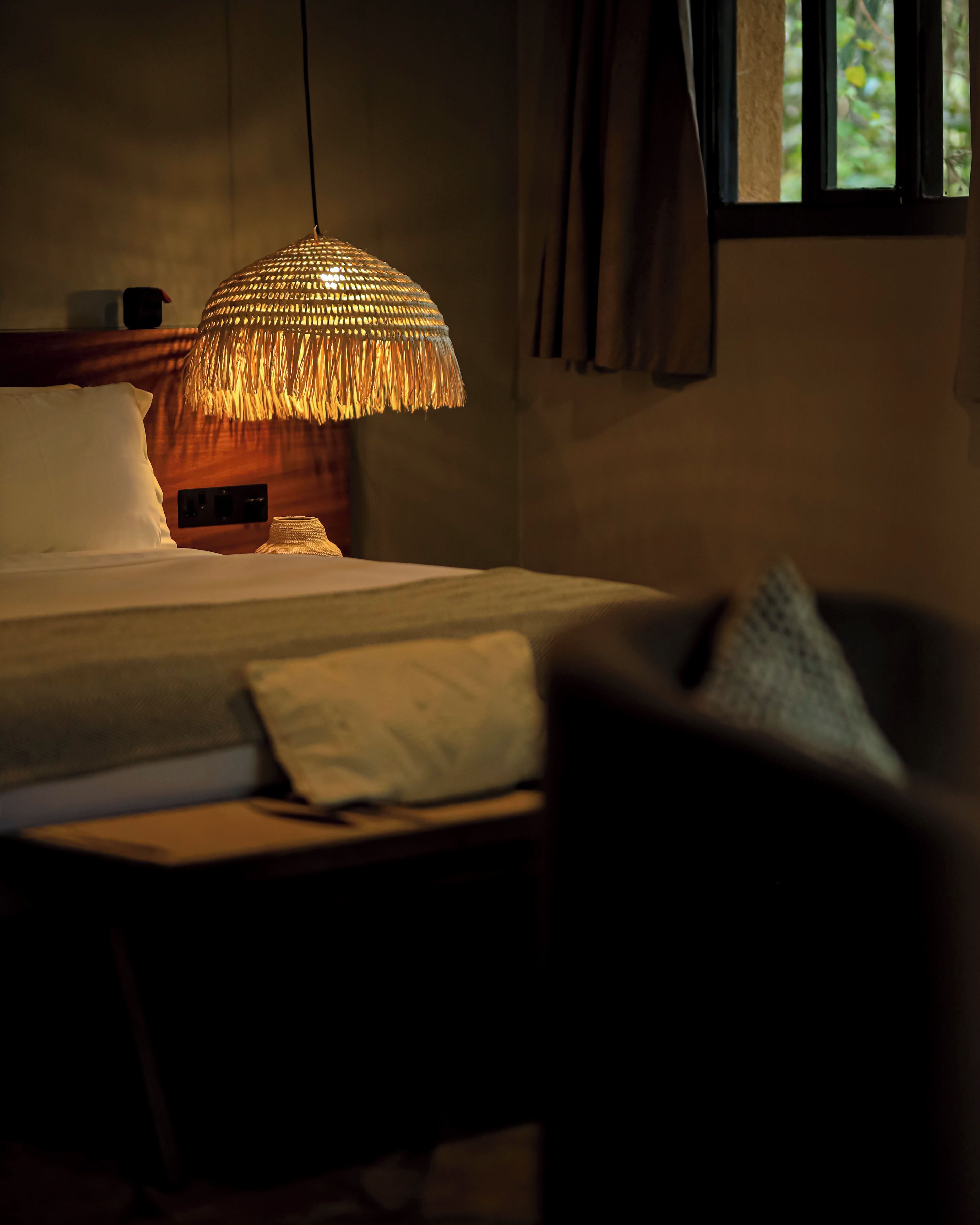 A dimly lit bedroom with a bed, a bedside table, a lamp, and a chair.