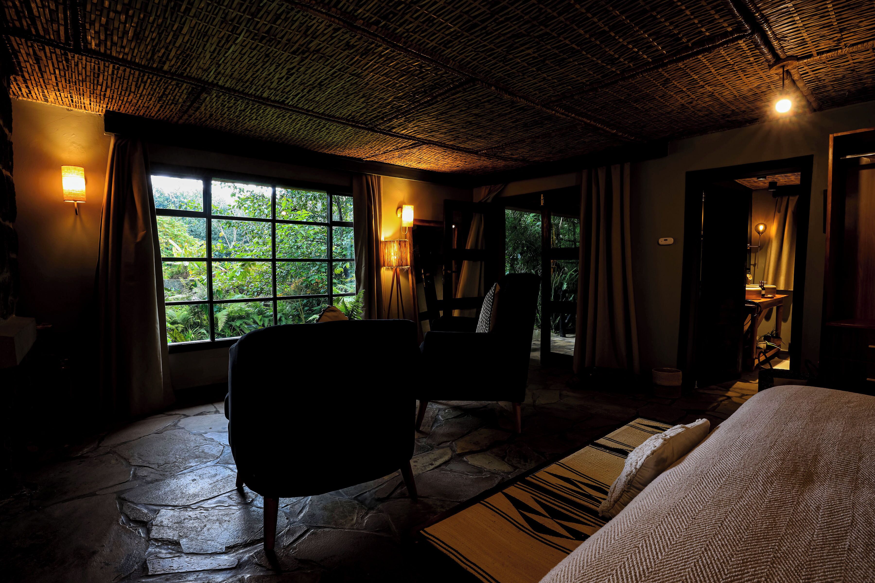 A room with a stone floor, a bed, a chair, and a view of greenery through the window.
