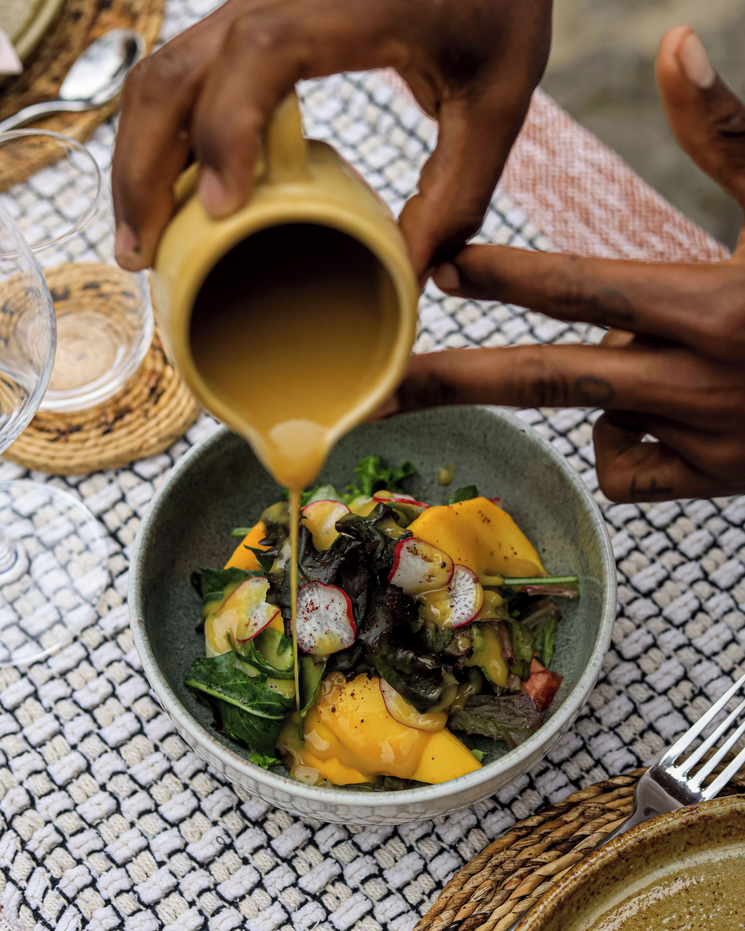A person pouring a liquid from a yellow cup into a bowl containing a salad with yellow fruit and greens.