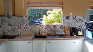 Una cocina con armarios blancos, una ventana con vista a árboles, un lavamanos y un hervidor.