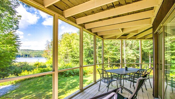 Un espacio al aire libre techado con una mesa y sillas, ventanas amplias y vistas a un lago y un bosque.