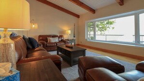 A spacious living room with a brown leather sofa, a wooden coffee table, and a large window providing a view of the outdoors. - Breathtaking 5 bedroom Waterfront Home With Hot Tub! (Port McNicoll)