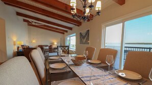 A dining room with a long table set for six, wooden beams on the ceiling, and a view of the sea through large windows. - Breathtaking 5 bedroom Waterfront Home With Hot Tub! (Port McNicoll)