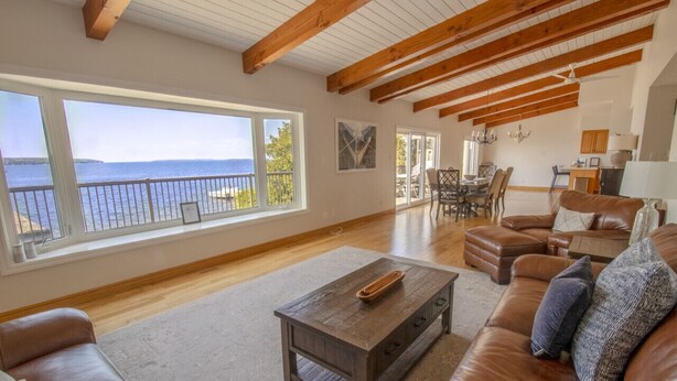 A spacious living room with a large window overlooking the sea, wooden beams on the ceiling, and a cozy seating area with leather sofas. - Breathtaking 5 bedroom Waterfront Home With Hot Tub! (Port McNicoll)