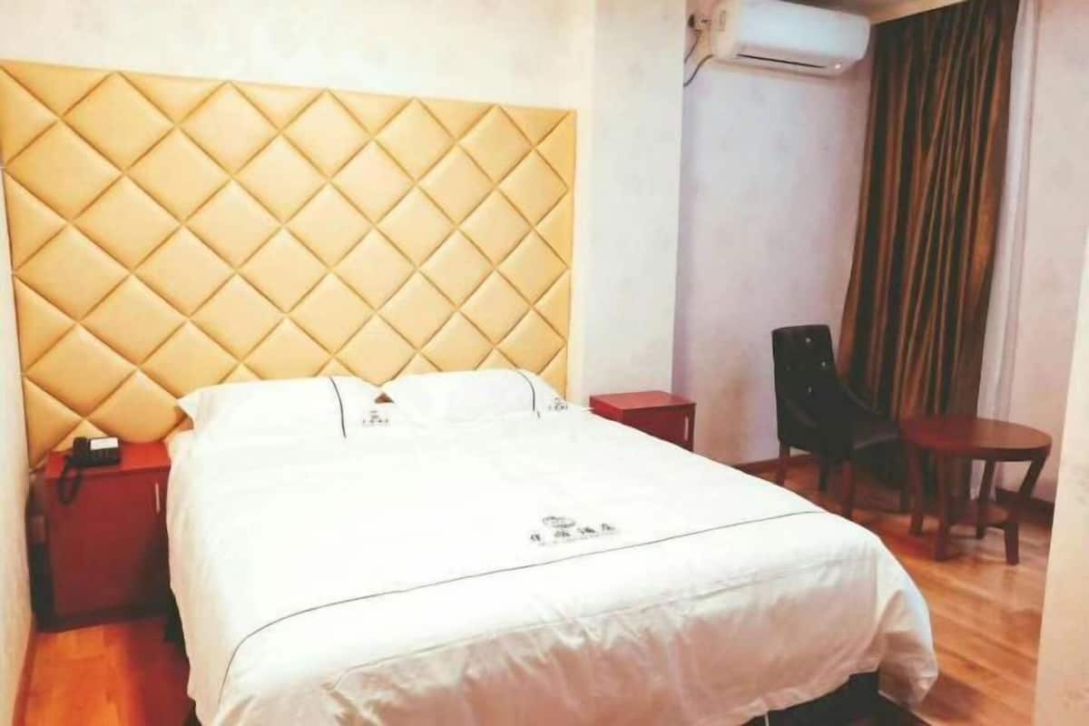 Habitación de hotel con cabecera acolchada, cama con ropa blanca, mesita de noche con teléfono, pequeña mesa con sillas y unidad de aire acondicionado en la pared.