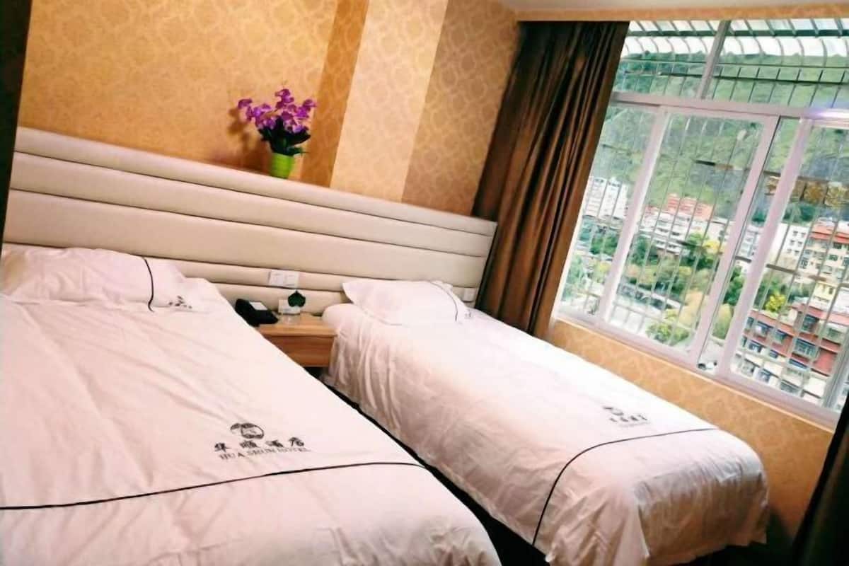 Habitación de hotel con dos camas, una mesita de noche, un florero con flores y una ventana con vistas a edificios.