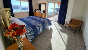 A bedroom with a bed, a wooden dresser, a chair, a vase with flowers, and a view of the sea through a balcony. - Sunrise Villa - Off-Grid Beach Villa w/ Best Views in Baja California! (Heroica Mulegé)