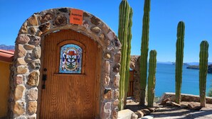 A stone archway with a wooden door and a stained glass window, leading to a cacti-lined path by a body of water. - Sunrise Villa - Off-Grid Beach Villa w/ Best Views in Baja California! (Heroica Mulegé)