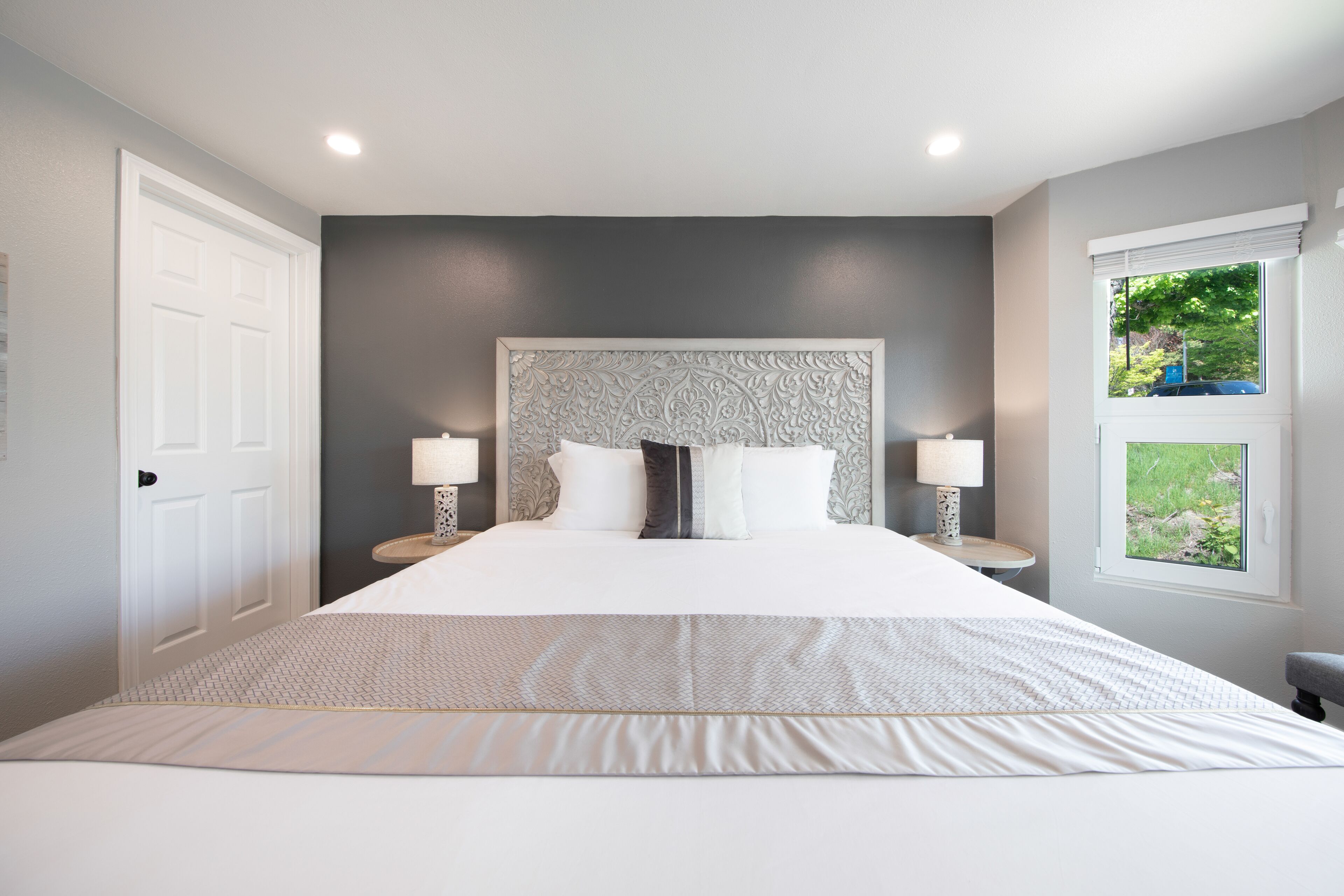 A large bed with a patterned headboard, two bedside lamps, and a window with a view of greenery.