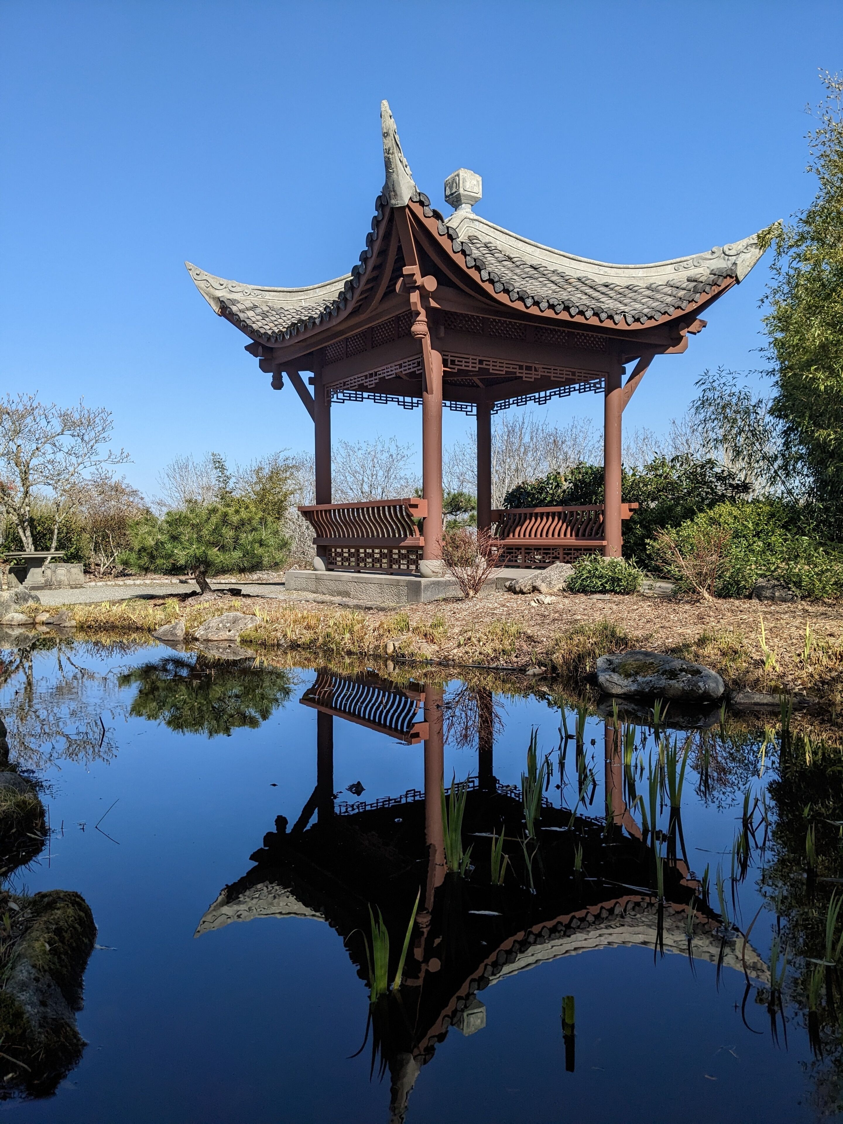 A traditional pavilion with a tiled roof and open sides, situated by a calm pond reflecting the structure.