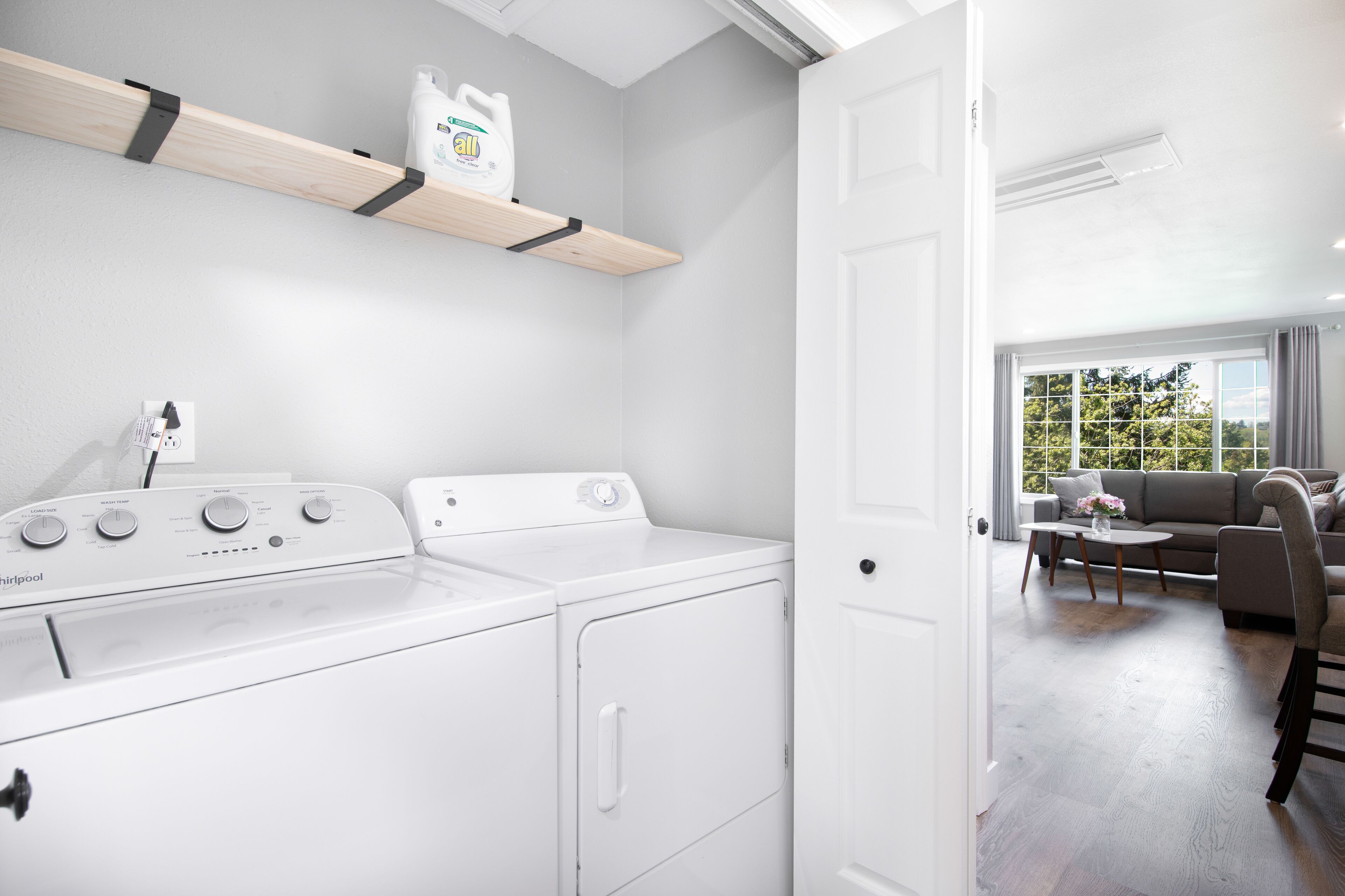 Un espace buanderie équipé d’un lave-linge et d’un sèche-linge Whirlpool, d’une étagère contenant de la lessive, et donnant sur un salon avec un canapé et une table.