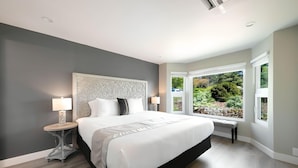 A modern bedroom with a large bed, two bedside tables with lamps, a bench, and a view of a garden through large windows. - High Point Guesthouse (Seattle)
